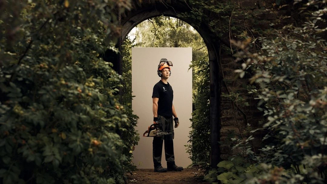 Man stands holding a chainsaw in front of a garden archway, wearing a protective helmet with ear protection, in an outdoor setting.