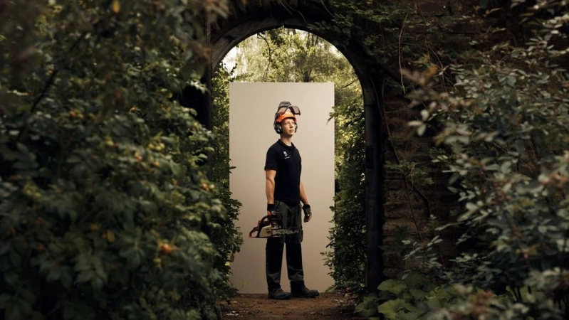 Man stands holding a chainsaw in front of a garden archway, wearing a protective helmet with ear protection, in an outdoor setting.