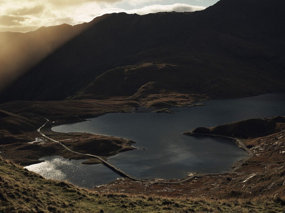 Scenic view of a dark blue lake surrounded by grassy hills and silhouetted mountains under a sunlit sky with visible sunbeams.