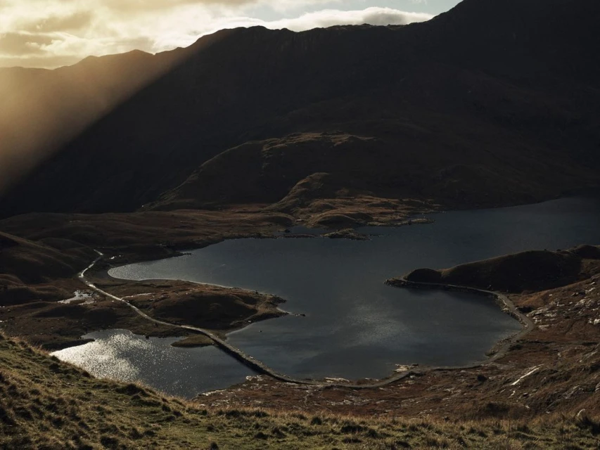 Scenic view of a dark blue lake surrounded by grassy hills and silhouetted mountains under a sunlit sky with visible sunbeams.