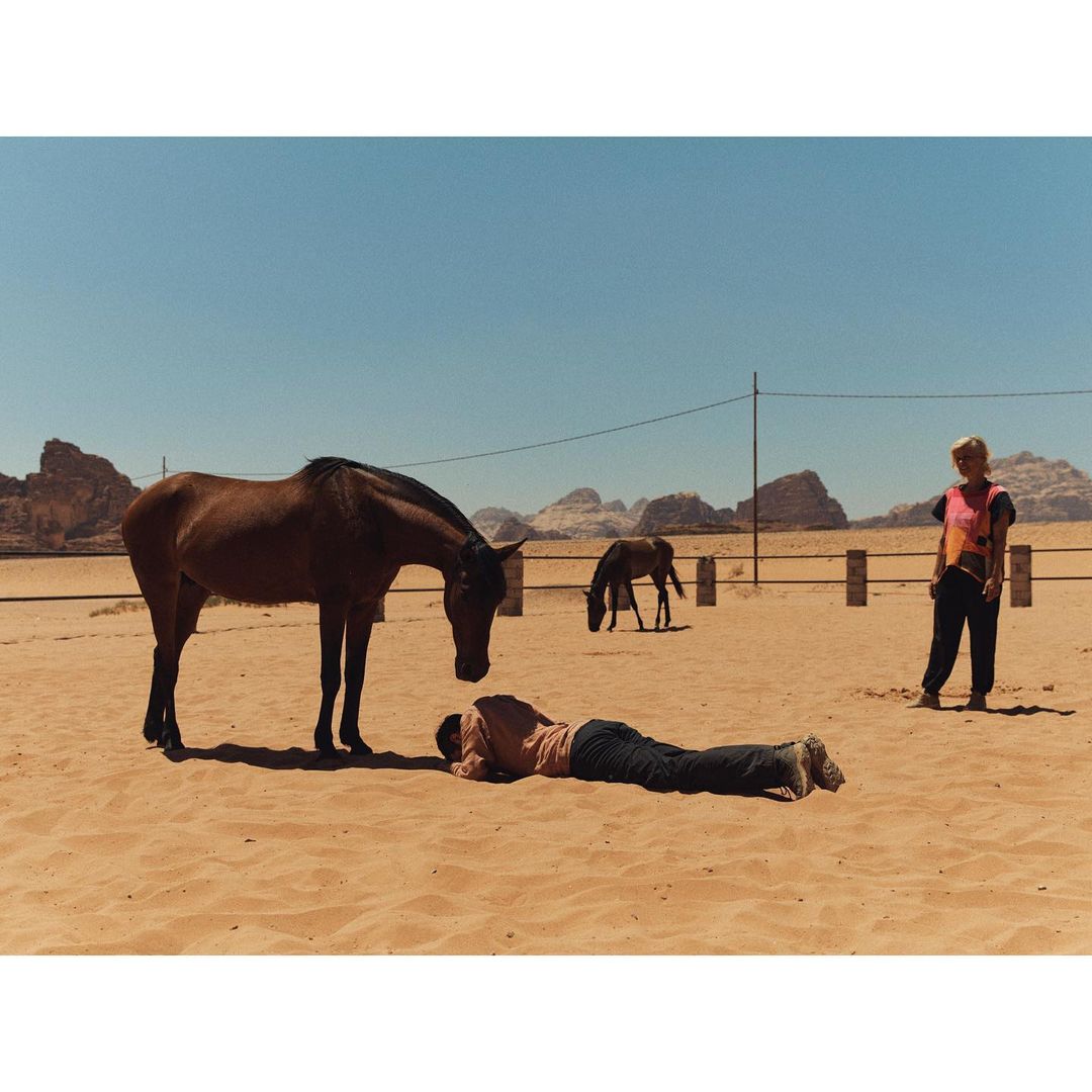 A brown horse stands over a man lying on the ground in a desert landscape, with a woman standing nearby and another horse in the background.