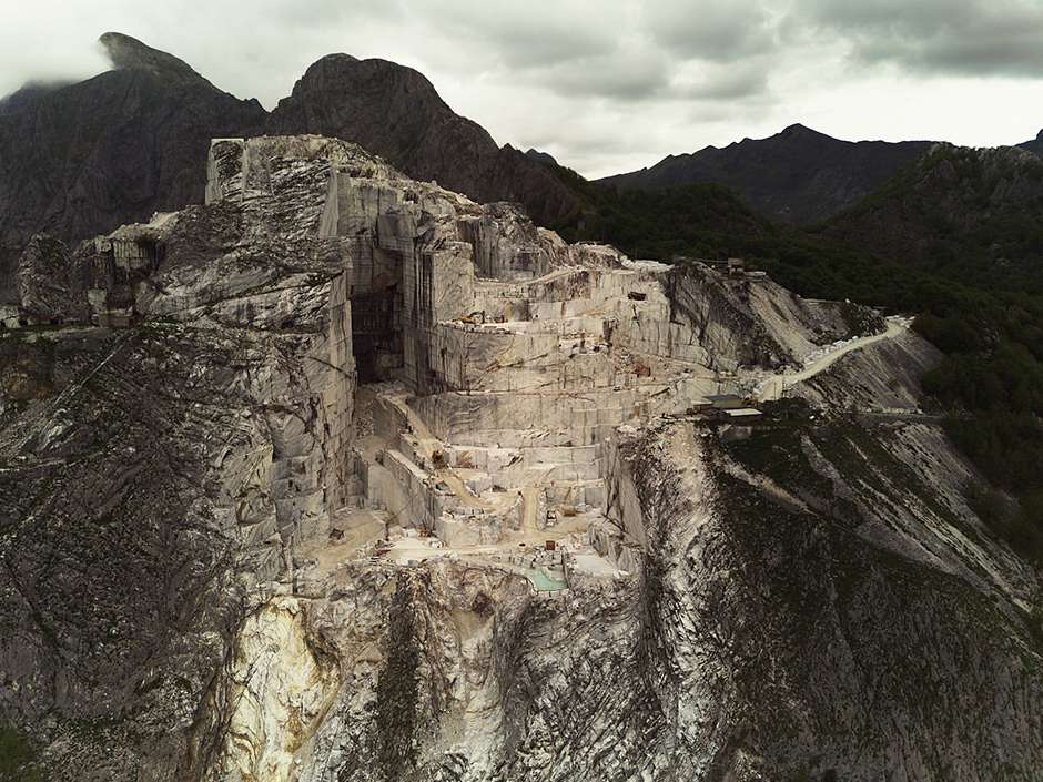 Aerial view of an open-pit Carrara marble quarry in Italy, featuring layered terraces, mining machinery, and surrounding mountains and forests.