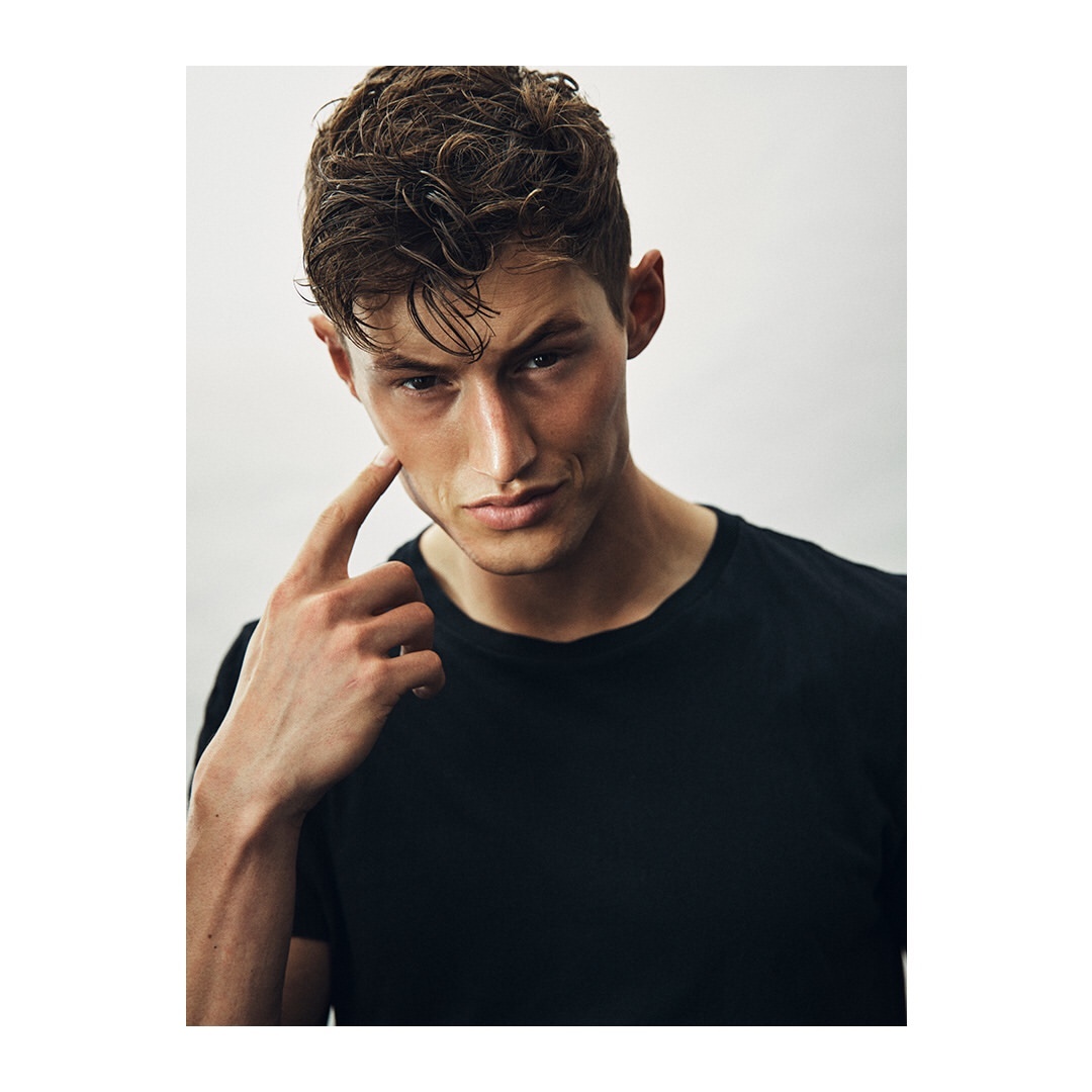 Portrait of a young adult male in a black t-shirt. He is touching his face and looking at the camera, set against a plain white background.