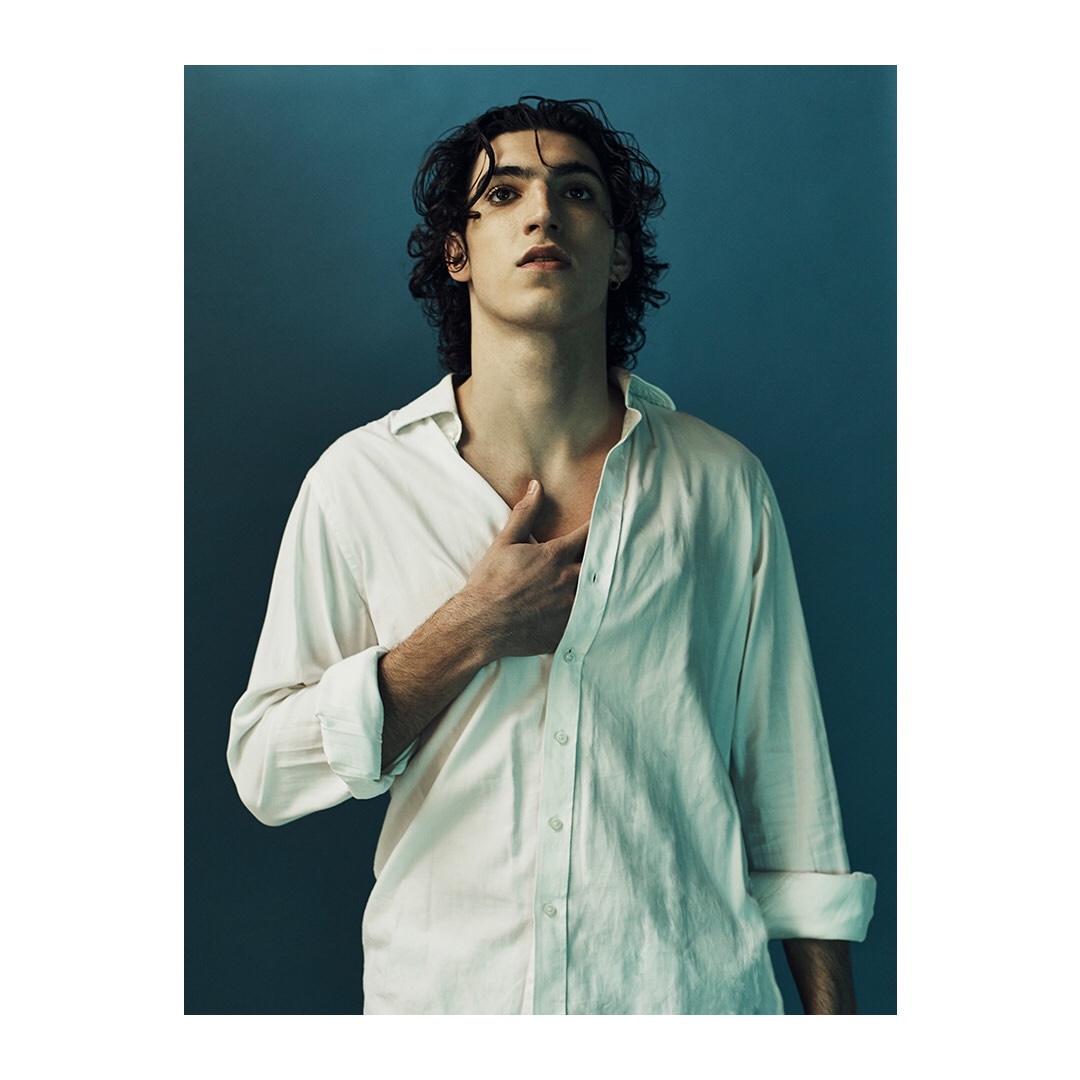 Portrait of a young adult man in a white button-down shirt against a teal background. He gazes upward with a hand near his chest.