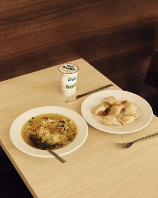 Overhead shot of two plates: one with soup and noodles, one with pierogi, and a container of kefir set on a light brown table.