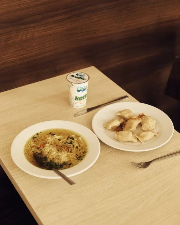 Overhead shot of two plates: one with soup and noodles, one with pierogi, and a container of kefir set on a light brown table.