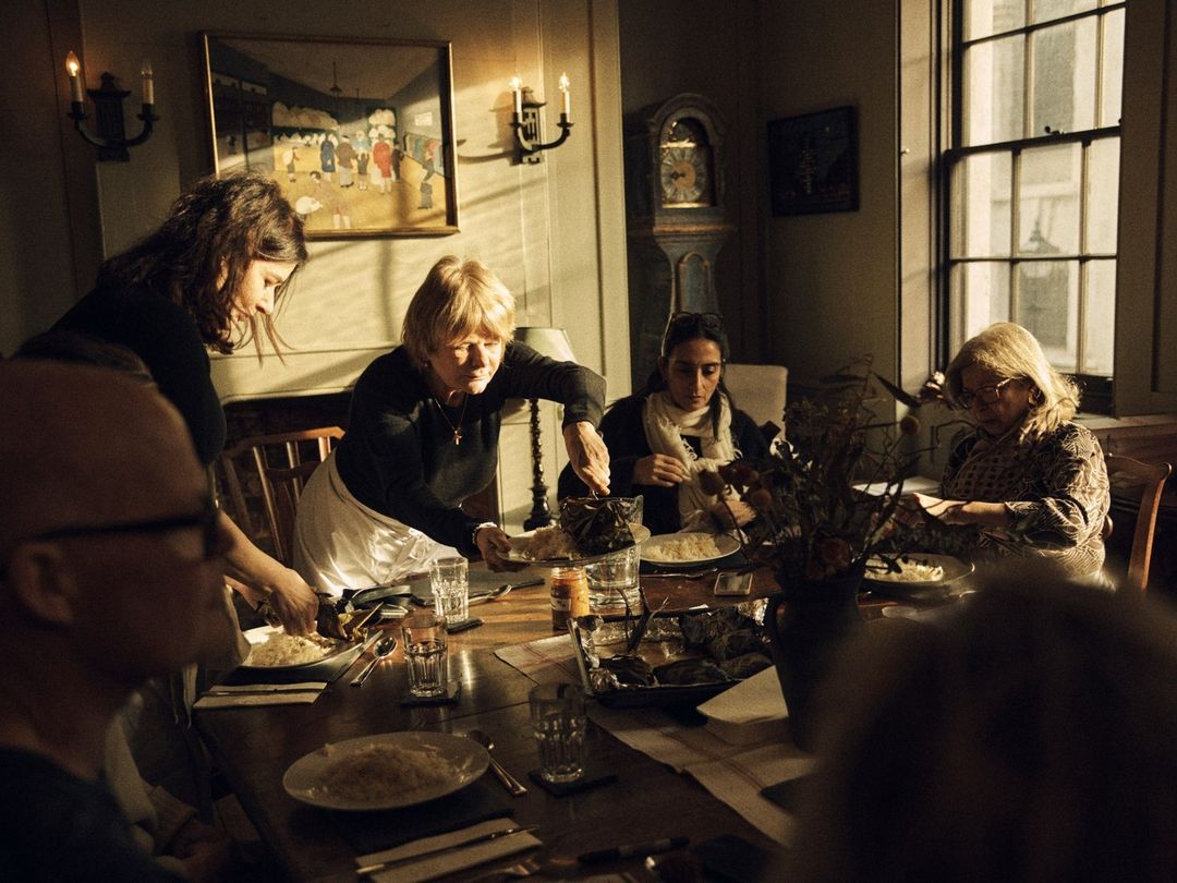 Candid photo of group of people gathered around a wooden table at an indoor dinner party in a warmly lit room with vintage decor.