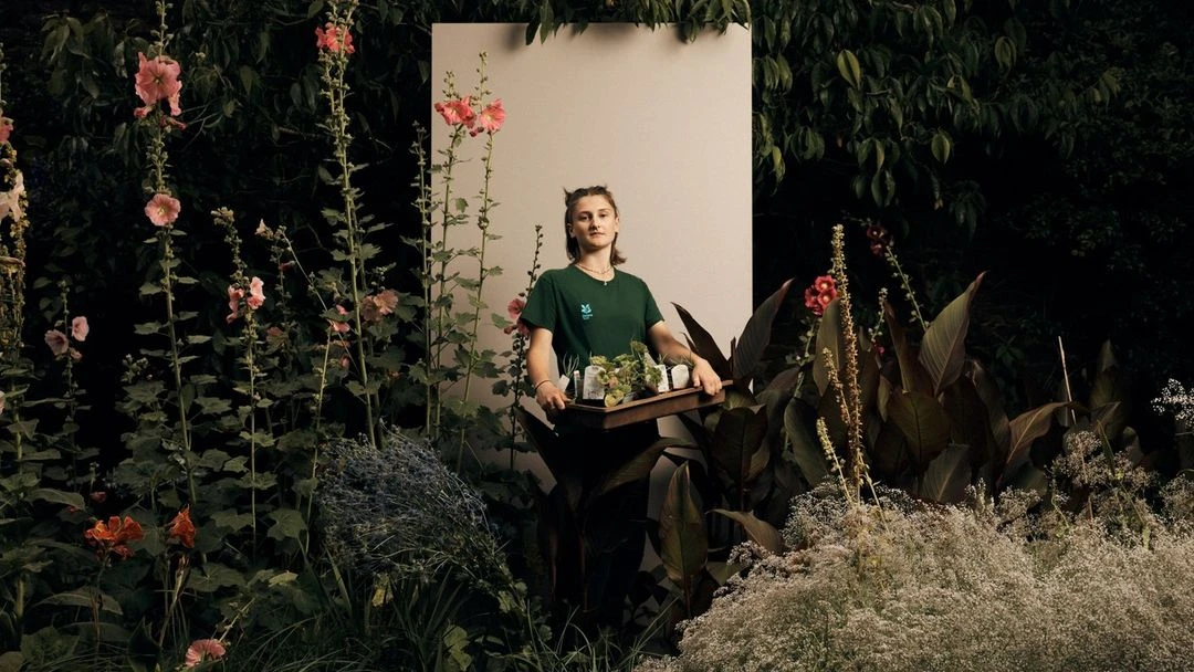 Woman with plant tray in lush garden: mid shot of a young woman holding plant seedlings, flowers, and foliage, neutral background.