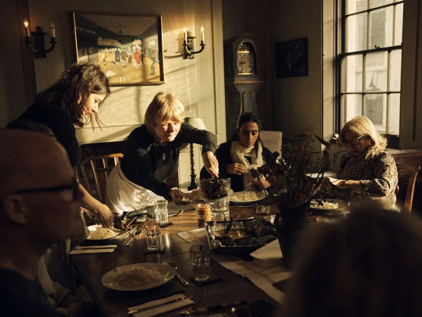 People gather around a wooden dining table in a warmly lit room, sharing food and conversation during a meal.