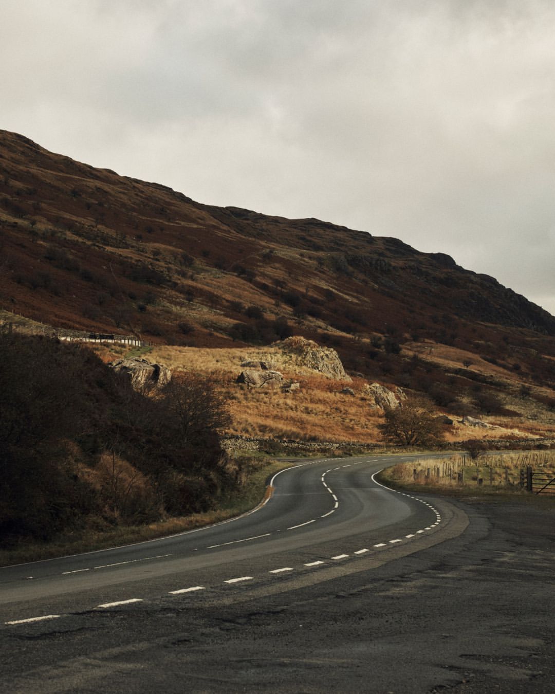 Winding rural road curves through a scenic landscape under an overcast sky with mountains and autumnal foliage.