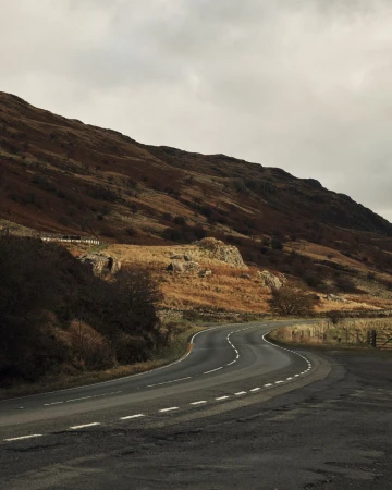 Winding rural road curves through a scenic landscape under an overcast sky with mountains and autumnal foliage.