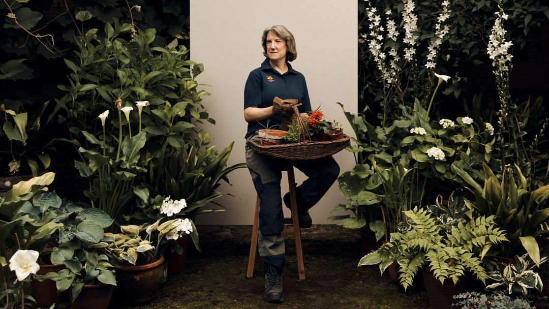 Adult woman sitting on a stool with a basket of potted plants surrounded by greenery in an outdoor garden setting.