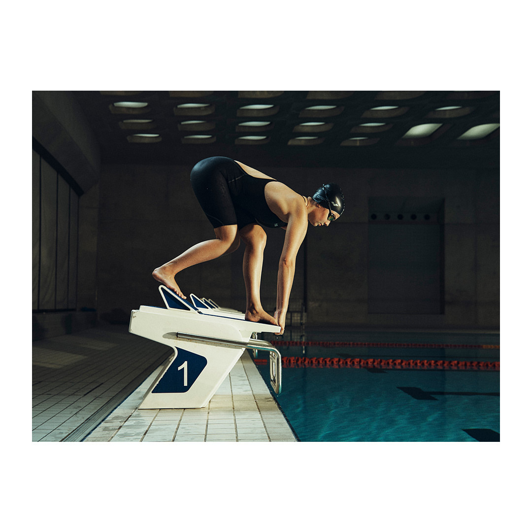 A female swimmer with a black cap and suit prepares to dive into an indoor pool from a white starting block with the number one on it.