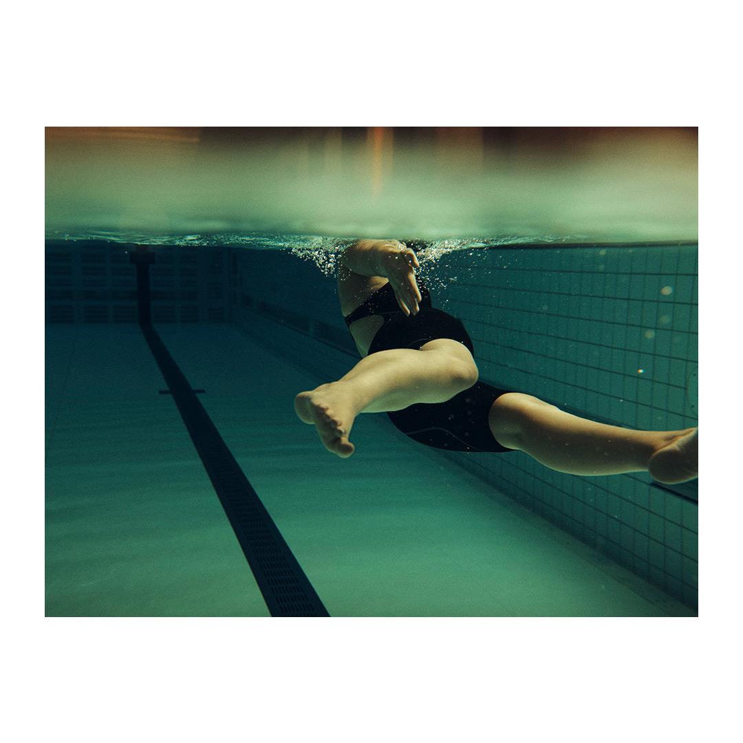 Underwater shot of a person swimming with a black swimsuit in a pool, showcasing the swimmer's form and the pool's lane markers.