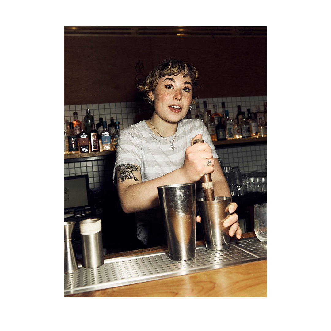 Adult female bartender in a striped t-shirt stands behind a wooden bar, making drinks with stainless steel shaker and barware.