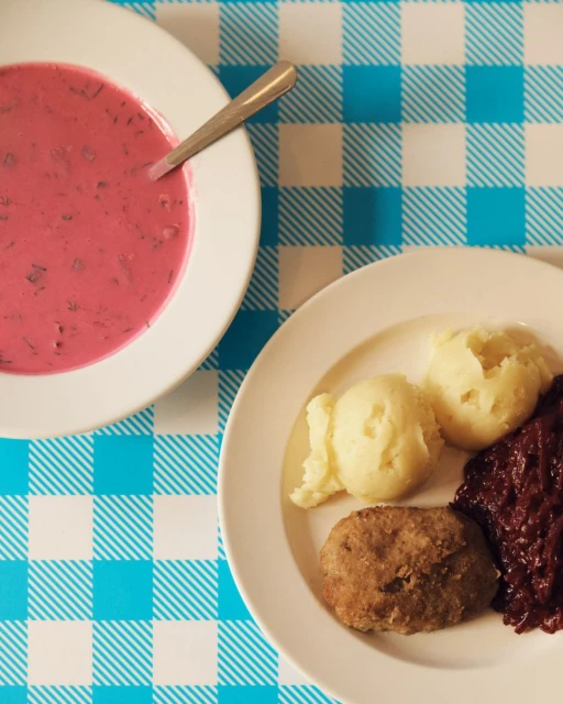 Overhead shot of a bowl of cold soup with a spoon and a plate of cutlet, potatoes, and red cabbage on a blue-and-white checkered tablecloth.
