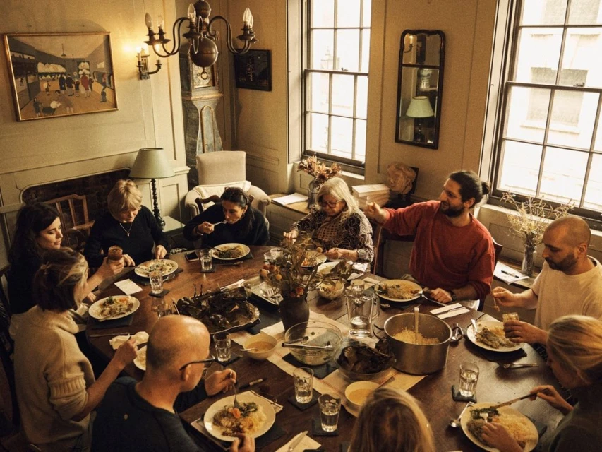 Large group of people of varying ages gathered around a wooden dining table set with plates of food in a warmly lit, traditional interior.