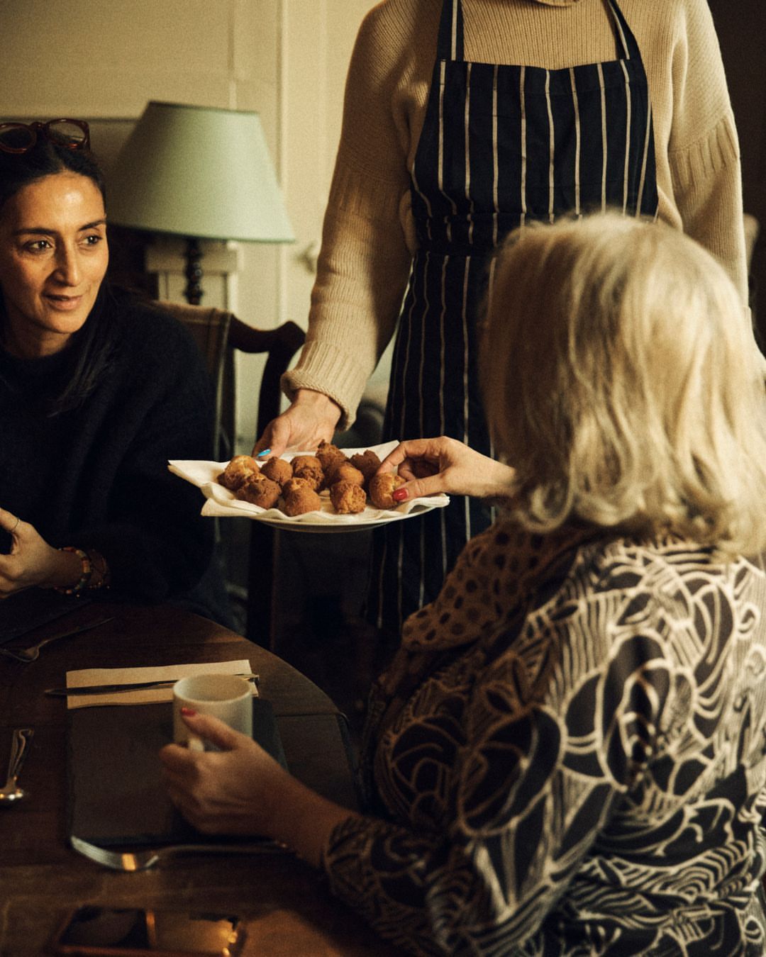 Three people sit around a table as a woman in an apron presents a platter of fried foods. The table is set in an indoor dining space.