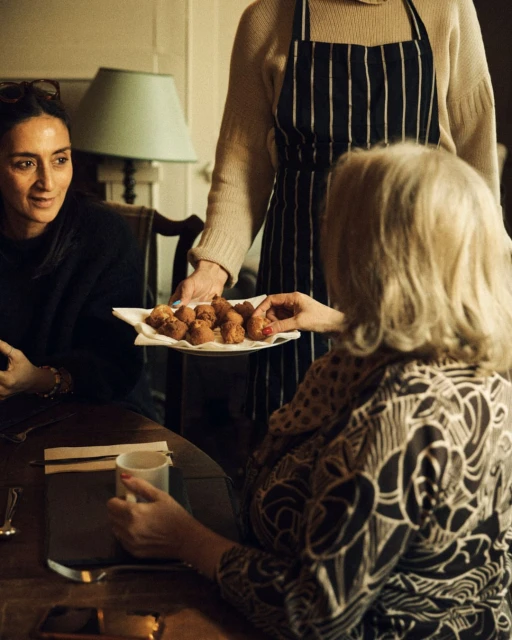 Three people sit around a table as a woman in an apron presents a platter of fried foods. The table is set in an indoor dining space.