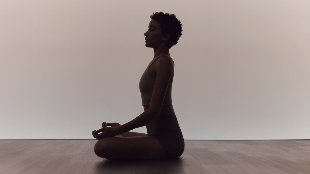 Adult woman in sportswear sits cross-legged in a meditation pose on a wooden floor against a light wall in a tranquil indoor setting.
