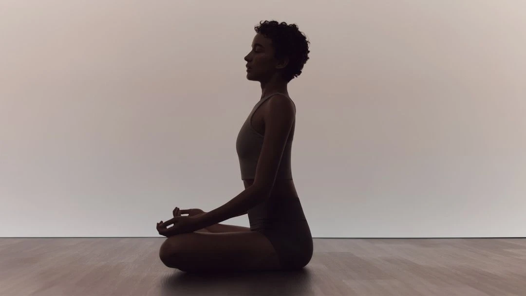 Adult woman in sportswear sits cross-legged in a meditation pose on a wooden floor against a light wall in a tranquil indoor setting.