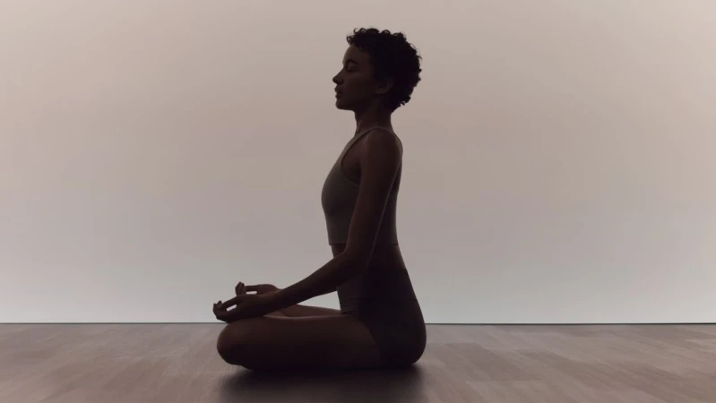 Adult woman in sportswear sits cross-legged in a meditation pose on a wooden floor against a light wall in a tranquil indoor setting.