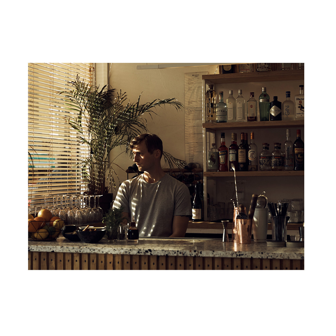 Bartender in a gray shirt stands behind a wooden bar with drinks and glassware, backlit near a window with a houseplant.