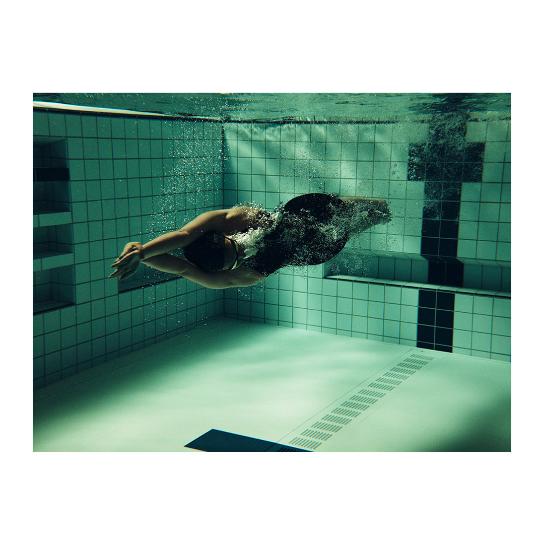 Underwater view of a swimmer in a dark swimsuit gliding through clear water in a tiled pool, showcasing fitness and aquatic sport.