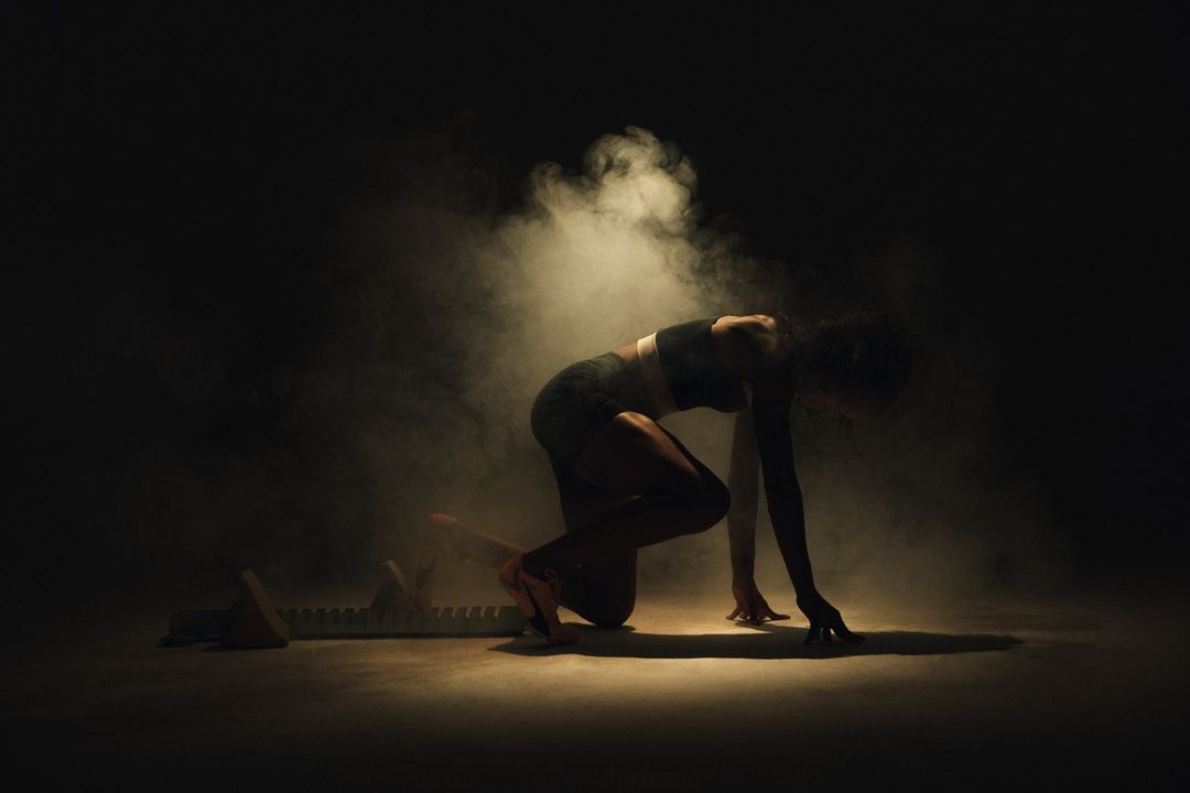 A female athlete is in a starting position on a track with starting blocks, set against a dark backdrop filled with atmospheric smoke.