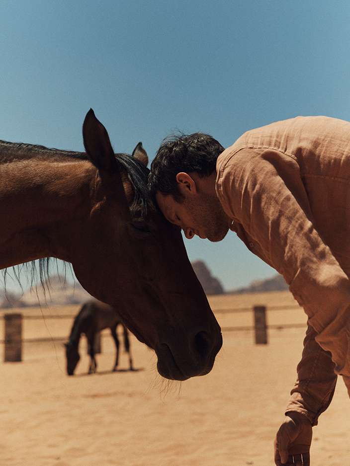 An adult male embraces a horse in a desert environment with a clear blue sky, showcasing a close bond between human and animal.