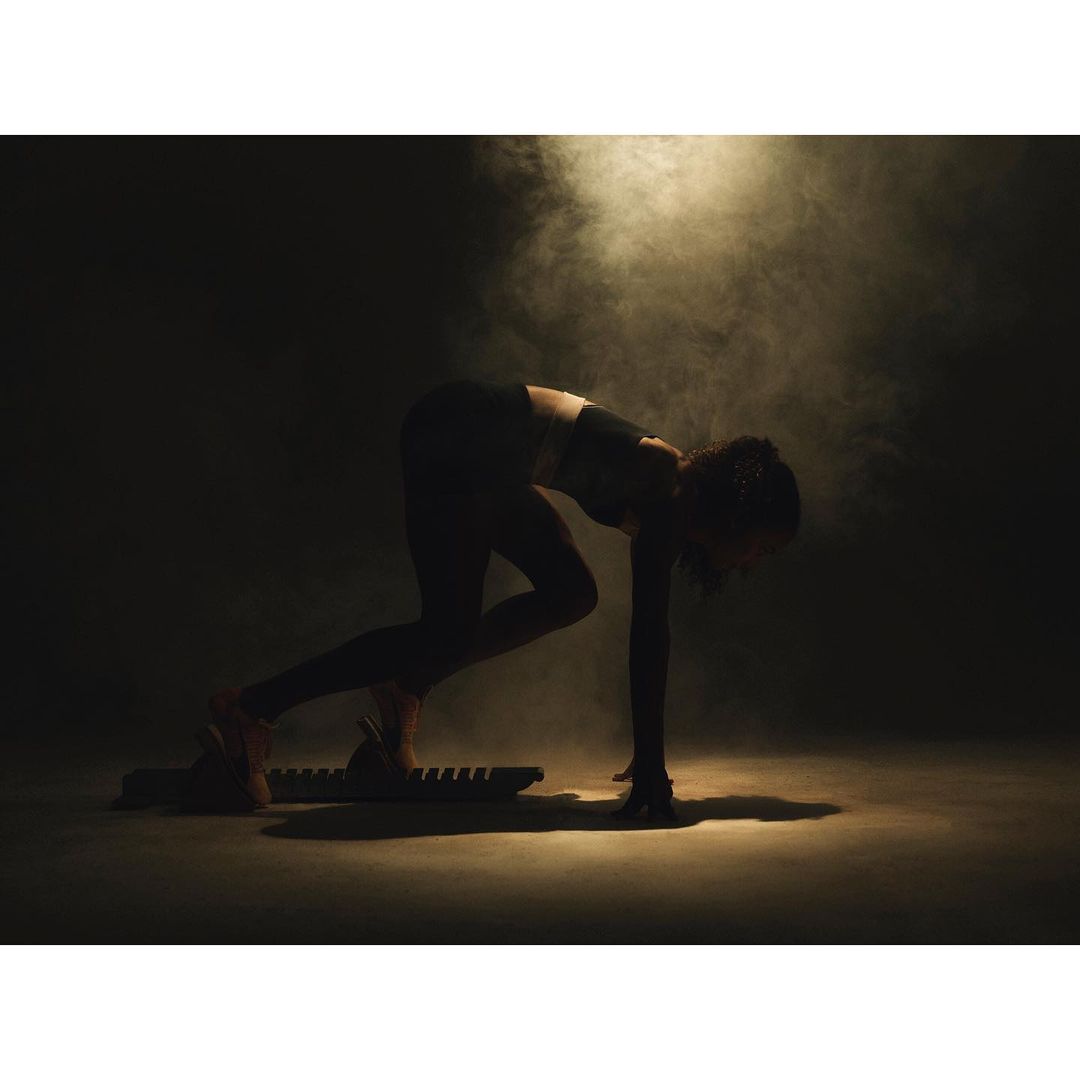 Female athlete in dark sportswear poses on a sled fitness board amidst smoky, moody lighting in a fitness setting.