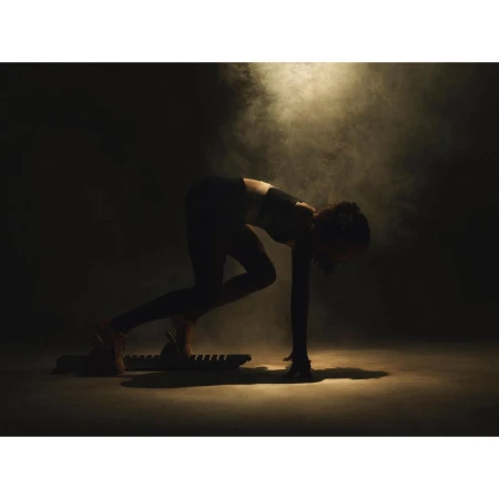 Female athlete in dark sportswear poses on a sled fitness board amidst smoky, moody lighting in a fitness setting.