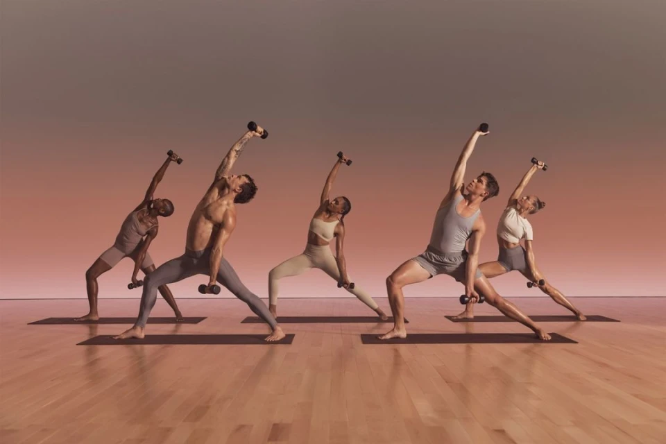 A group of five diverse adults are in a yoga studio, balancing in matching sportswear while holding dumbbells, with a clean backdrop.