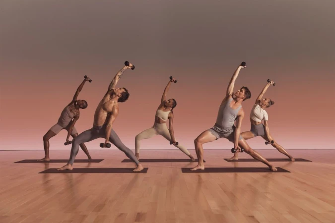 A group of five diverse adults are in a yoga studio, balancing in matching sportswear while holding dumbbells, with a clean backdrop.
