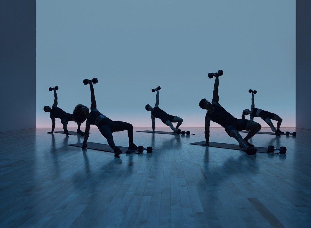 Group fitness class with five people doing a side plank dumbbell exercise in a blue-lit studio with wood floors and a minimalist background.
