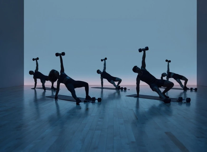 Group fitness class with five people doing a side plank dumbbell exercise in a blue-lit studio with wood floors and a minimalist background.