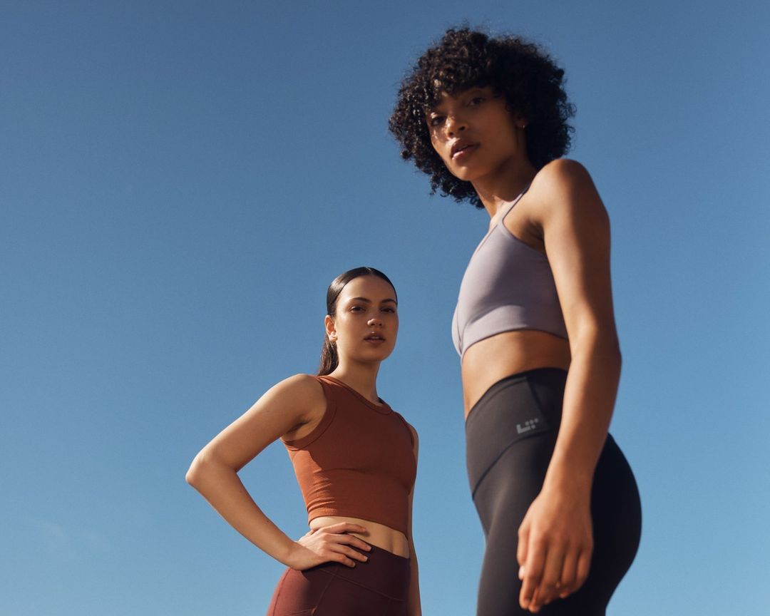 Two athletic women in sports bras and leggings pose against a clear blue sky, shot from a low angle emphasizing their active lifestyle.
