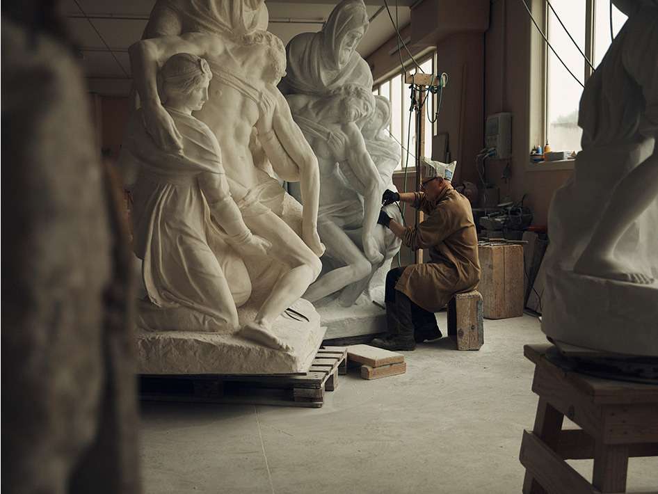 Sculptor working on a large marble sculpture in a studio, shaping the stone with hand tools, in a workspace with other sculptures.