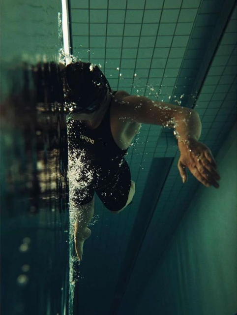 Underwater shot of a swimmer in a black swimsuit with the Arena logo, swimming in a pool with green tiled walls.
