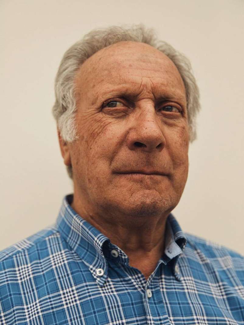 Head and shoulders portrait of an elderly adult man with grey hair and a blue plaid shirt against a plain off-white background.