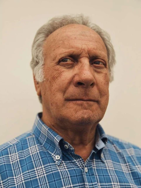 Head and shoulders portrait of an elderly adult man with grey hair and a blue plaid shirt against a plain off-white background.
