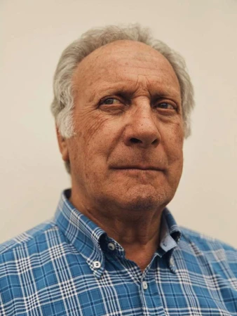 Head and shoulders portrait of an elderly adult man with grey hair and a blue plaid shirt against a plain off-white background.
