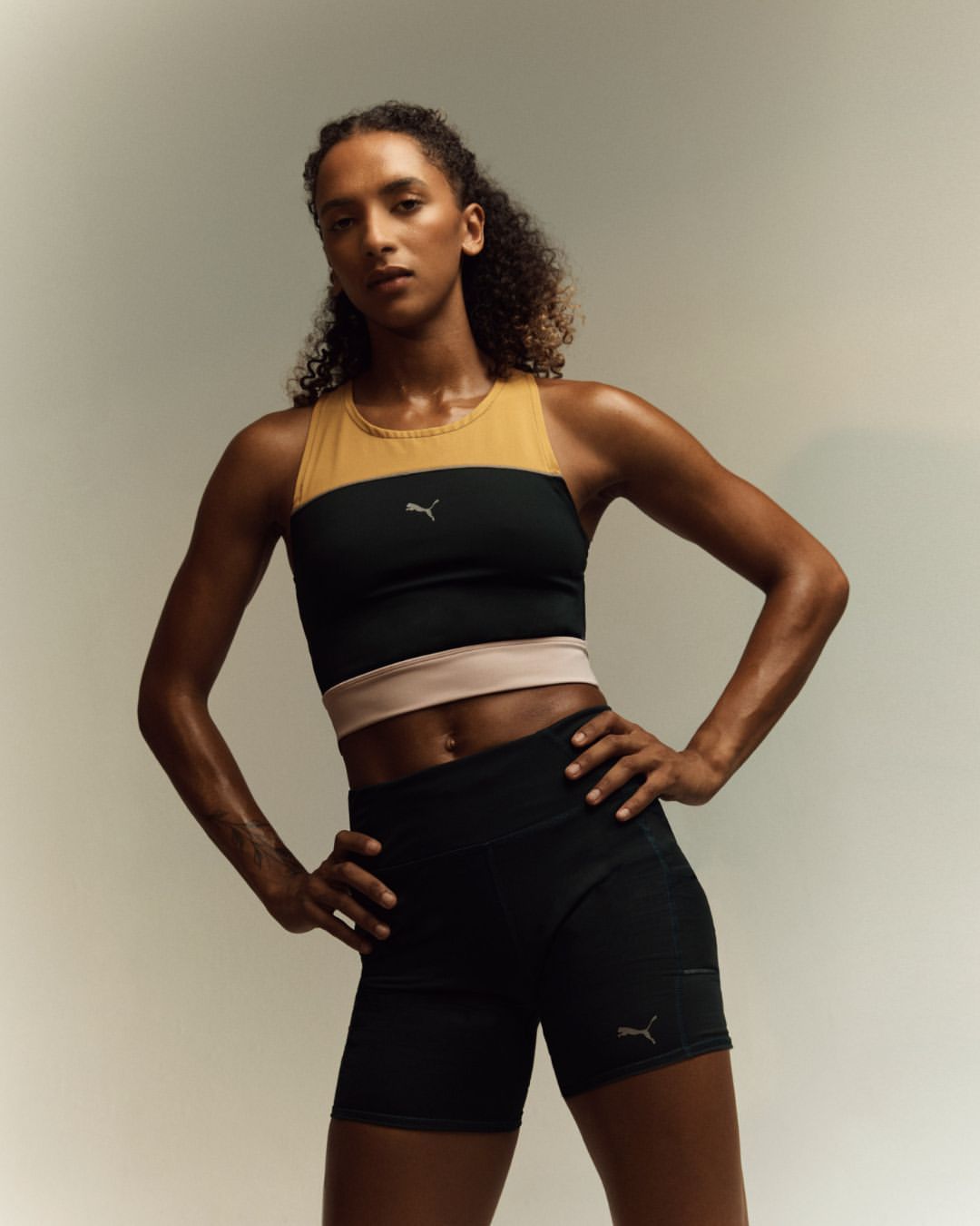 Athletic woman in sportswear poses confidently in a studio. She wears a color-blocked top and shorts with a neutral background and bright lighting.