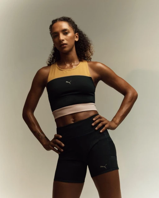 Athletic woman in sportswear poses confidently in a studio. She wears a color-blocked top and shorts with a neutral background and bright lighting.