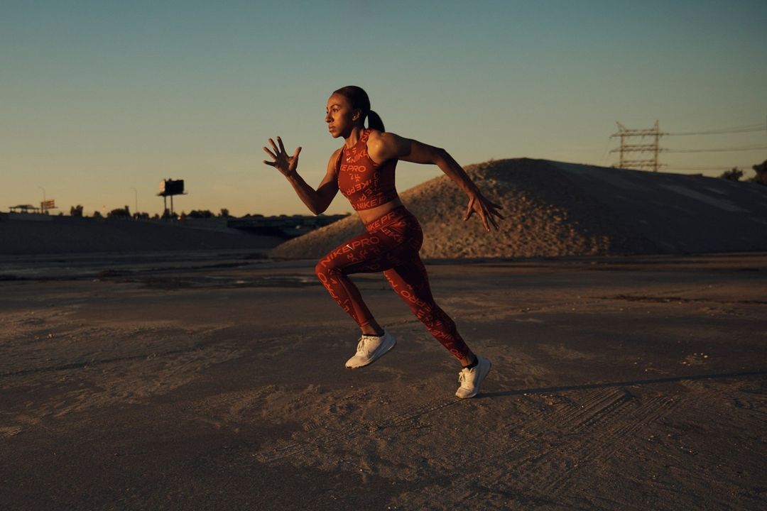 Athletic woman sprints across a sandy landscape at sunset, dressed in red Nike Pro athletic wear and white sneakers.