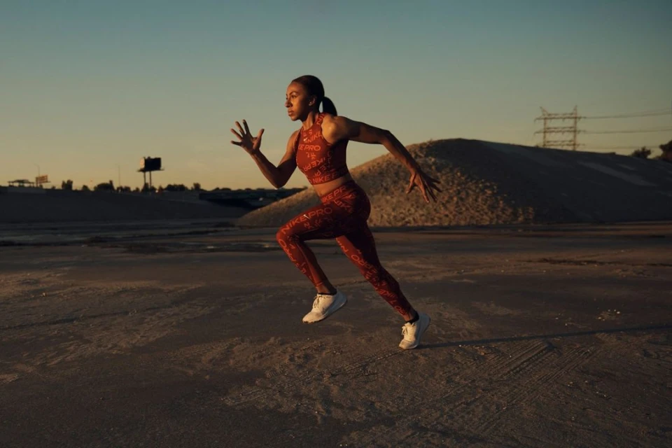 Athletic woman sprints across a sandy landscape at sunset, dressed in red Nike Pro athletic wear and white sneakers.