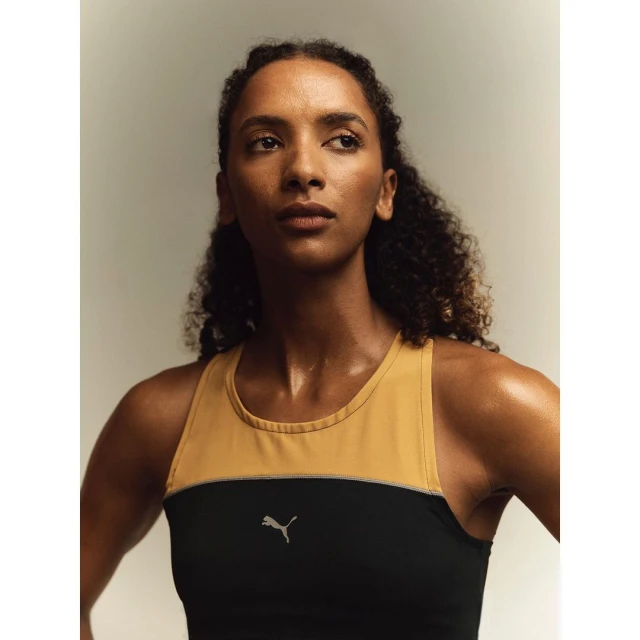Adult woman with curly hair poses in a color-blocked athletic top with the Puma logo, captured in a medium shot against a neutral backdrop.