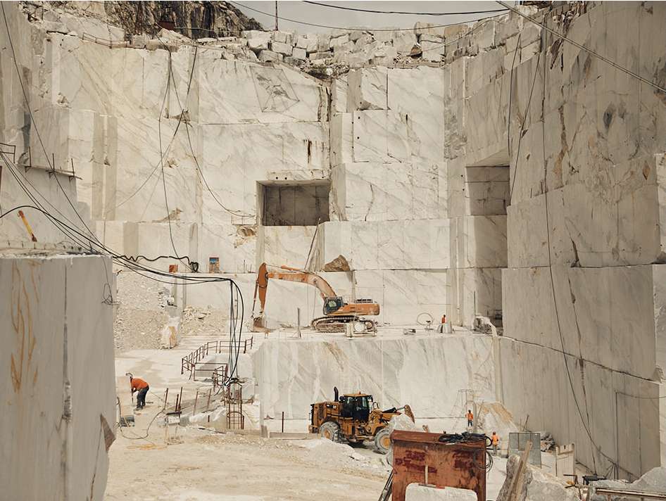 Large-scale marble quarry with workers, heavy machinery, and excavation equipment against massive cut marble walls in an outdoor setting.