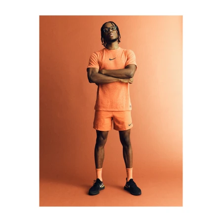 Black man with braided hair wearing an orange athletic t-shirt and shorts with arms crossed