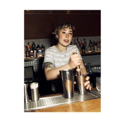Female bartender in striped t-shirt muddling a cocktail behind a bar counter in London by Dave Imms